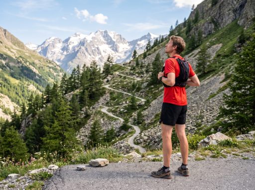 Coureur en tenue de trail au bout de la route goudronnée, face à un sentier en lacets qui monte vers les sommets des Alpes
